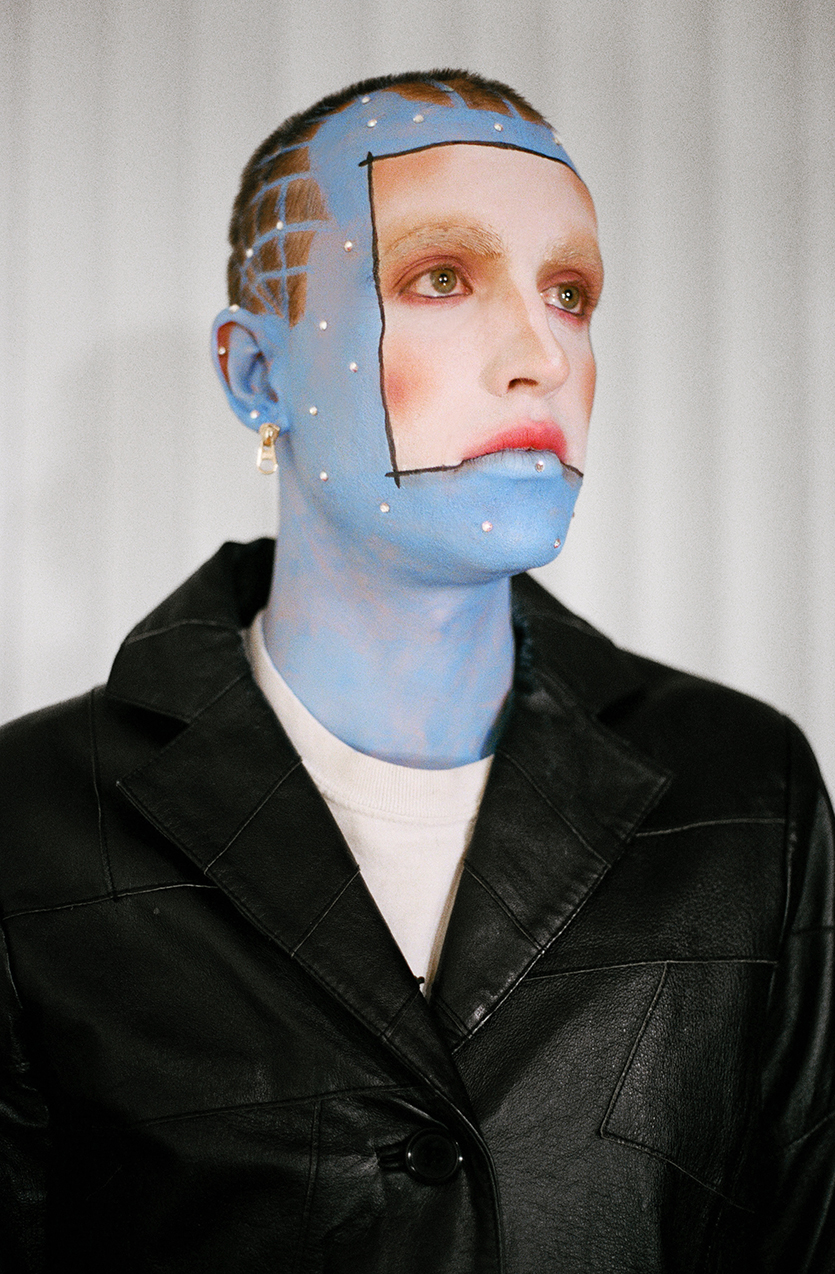 Close up photo of a model looking beyond the cmaera with blue skin and and a white square in the center of their face wearing a black leather jacket in front of a white background
